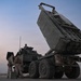 The image shows a multiple launch rocket system (MLRS) mounted on a heavy military truck with its launcher pod elevated at an angle. Several military personnel in camouflage uniforms are gathered around the vehicle, apparently conducting operations or maintenance. The scene appears to take place on a paved surface during dusk or dawn conditions.