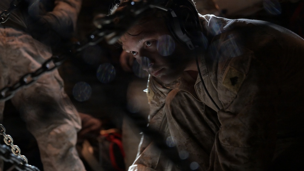 A military service member wearing headset communications equipment and desert camouflage uniform is working in a dimly lit environment, likely the cargo hold of a military aircraft. The person appears focused and is leaning forward, working with chains and cargo securing equipment. The image features dramatic bokeh light effects overlaid on the scene.