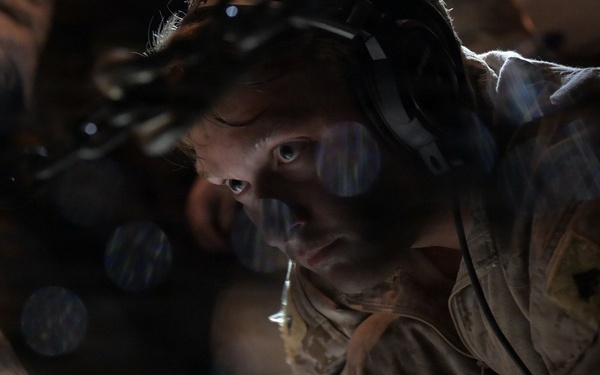 A military service member wearing headset communications equipment and desert camouflage uniform is working in a dimly lit environment, likely the cargo hold of a military aircraft. The person appears focused and is leaning forward, working with chains and cargo securing equipment. The image features dramatic bokeh light effects overlaid on the scene.