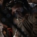 A military service member wearing headset communications equipment and desert camouflage uniform is working in a dimly lit environment, likely the cargo hold of a military aircraft. The person appears focused and is leaning forward, working with chains and cargo securing equipment. The image features dramatic bokeh light effects overlaid on the scene.