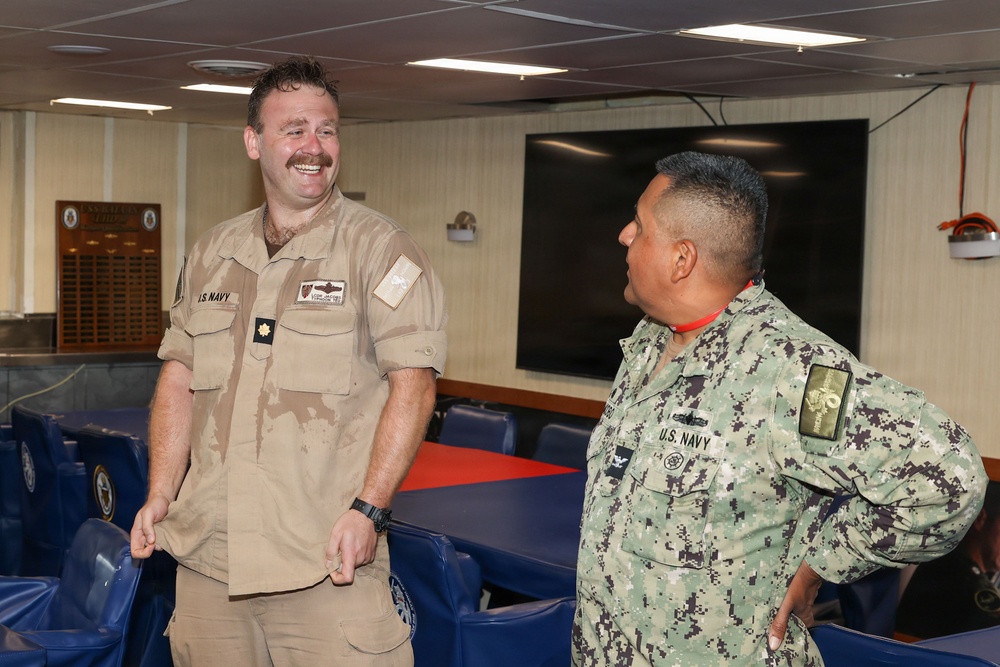 Two U.S. Navy personnel are engaged in a casual conversation inside what appears to be a shipboard conference or briefing room. The individual on the left is wearing tan/khaki working uniform with a mustache and is laughing, while the person on the right is wearing the Navy Working Uniform (NWU) digital camouflage pattern. Both have 'U.S. Navy' patches visible on their uniforms.