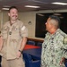 Two U.S. Navy personnel are engaged in a casual conversation inside what appears to be a shipboard conference or briefing room. The individual on the left is wearing tan/khaki working uniform with a mustache and is laughing, while the person on the right is wearing the Navy Working Uniform (NWU) digital camouflage pattern. Both have 'U.S. Navy' patches visible on their uniforms.