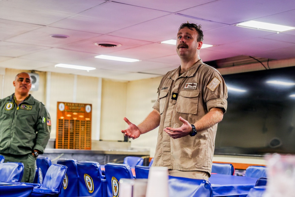 Two military personnel are present in what appears to be a briefing room or mess hall aboard a naval vessel or installation. The central figure, wearing a U.S. Navy khaki uniform with a gold oak leaf insignia (indicating the rank of Lieutenant Commander or Major), is actively speaking and gesturing with his hands. A second individual in a green flight suit stands in the background observing.