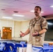 Two military personnel are present in what appears to be a briefing room or mess hall aboard a naval vessel or installation. The central figure, wearing a U.S. Navy khaki uniform with a gold oak leaf insignia (indicating the rank of Lieutenant Commander or Major), is actively speaking and gesturing with his hands. A second individual in a green flight suit stands in the background observing.