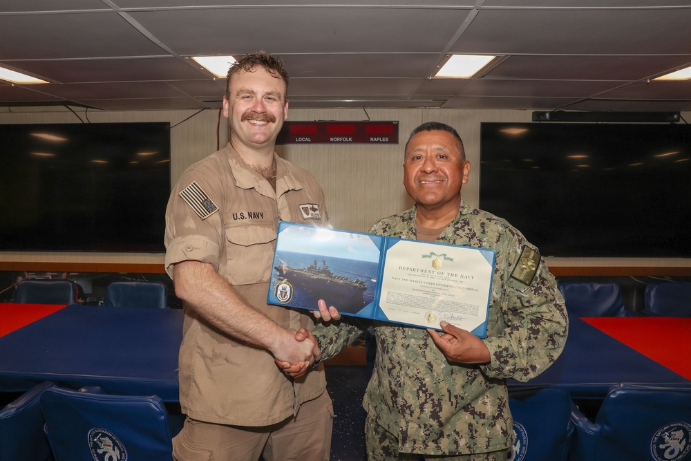 Two U.S. Navy personnel are posing together while holding an open award certificate from the Department of the Navy. The certificate appears to be a Navy and Marine Corps Commendation Medal citation. The ceremony is taking place in what appears to be a shipboard briefing or conference room.