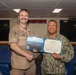 Two U.S. Navy personnel are posing together while holding an open award certificate from the Department of the Navy. The certificate appears to be a Navy and Marine Corps Commendation Medal citation. The ceremony is taking place in what appears to be a shipboard briefing or conference room.