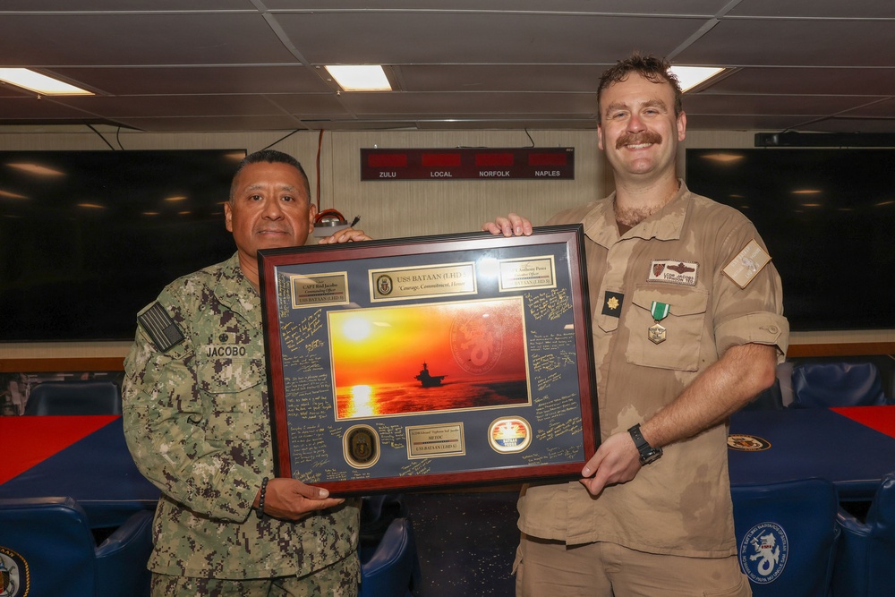Two military personnel are posing together while holding a framed commemorative plaque/shadow box for the USS Bataan (LHD-5). The individual on the left, identified by his name tape as 'Jacobo,' is wearing Navy Working Uniform (NWU) digital camouflage, while the individual on the right is wearing a tan/khaki uniform with a medal and rank insignia. The setting appears to be aboard a naval vessel, indicated by the digital clock display showing time zones (Zulu, Local, Norfolk, Naples) in the background.