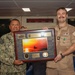 Two military personnel are posing together while holding a framed commemorative plaque/shadow box for the USS Bataan (LHD-5). The individual on the left, identified by his name tape as 'Jacobo,' is wearing Navy Working Uniform (NWU) digital camouflage, while the individual on the right is wearing a tan/khaki uniform with a medal and rank insignia. The setting appears to be aboard a naval vessel, indicated by the digital clock display showing time zones (Zulu, Local, Norfolk, Naples) in the background.