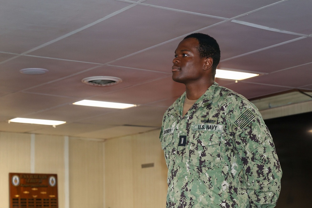 A U.S. Navy service member wearing the Navy Working Uniform (NWU) Type III in woodland digital camouflage pattern stands at attention or in a formal posture indoors. The uniform displays 'U.S. NAVY' chest tape, a name tape reading 'TURNER', an American flag patch on the right sleeve, and what appears to be an Ensign (O-1) rank insignia. The setting appears to be a military facility or command building.