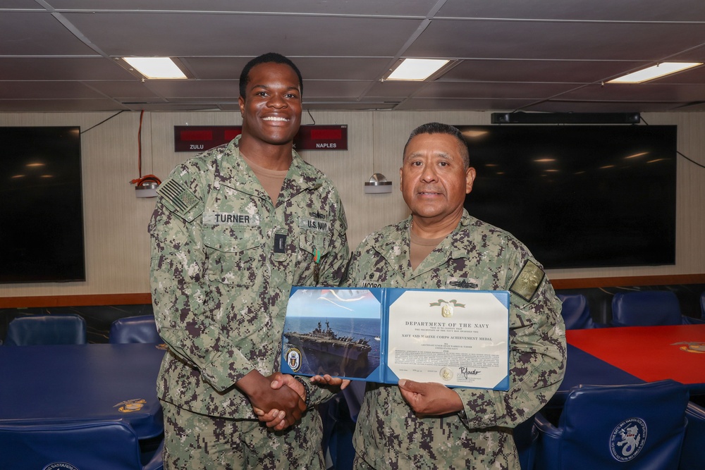 Two U.S. Navy personnel are posing together while holding a Department of the Navy certificate for the Navy and Marine Corps Achievement Medal. The taller sailor on the left, identified by name tape as 'TURNER,' is shaking hands with the shorter senior sailor on the right. The ceremony appears to be taking place in a shipboard briefing or conference room.