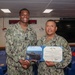 Two U.S. Navy personnel are posing together while holding a Department of the Navy certificate for the Navy and Marine Corps Achievement Medal. The taller sailor on the left, identified by name tape as 'TURNER,' is shaking hands with the shorter senior sailor on the right. The ceremony appears to be taking place in a shipboard briefing or conference room.