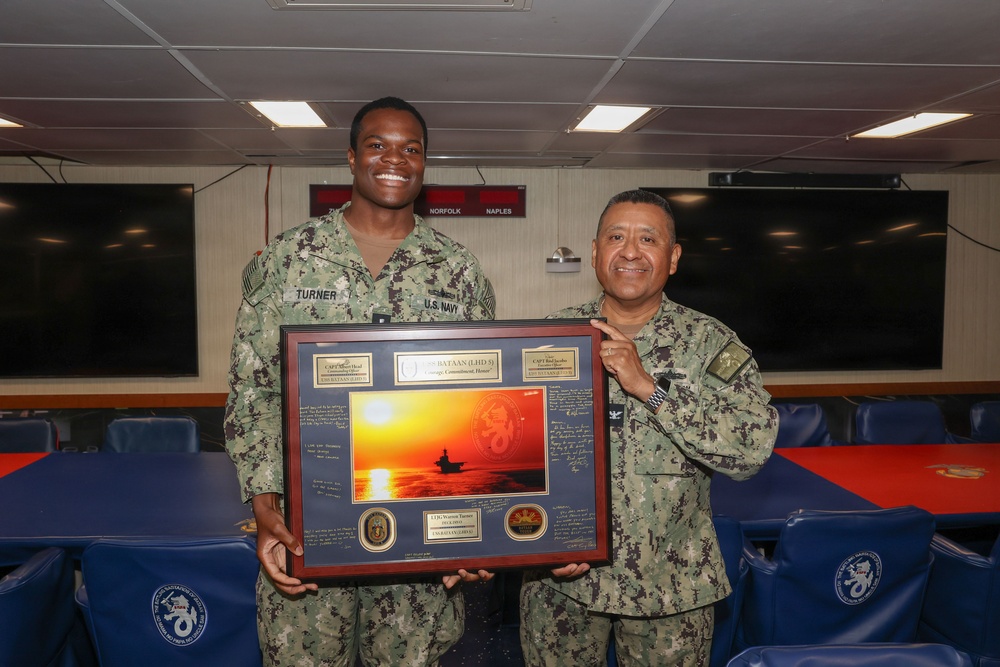 Two U.S. Navy personnel in digital camouflage uniforms are posing together in what appears to be a shipboard conference or briefing room. They are holding a framed commemorative plaque/shadow box featuring a sunset photograph and what appears to be USS Bataan (LHD-5) insignia. The occasion appears to be a formal recognition or farewell ceremony.