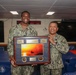Two U.S. Navy personnel in digital camouflage uniforms are posing together in what appears to be a shipboard conference or briefing room. They are holding a framed commemorative plaque/shadow box featuring a sunset photograph and what appears to be USS Bataan (LHD-5) insignia. The occasion appears to be a formal recognition or farewell ceremony.