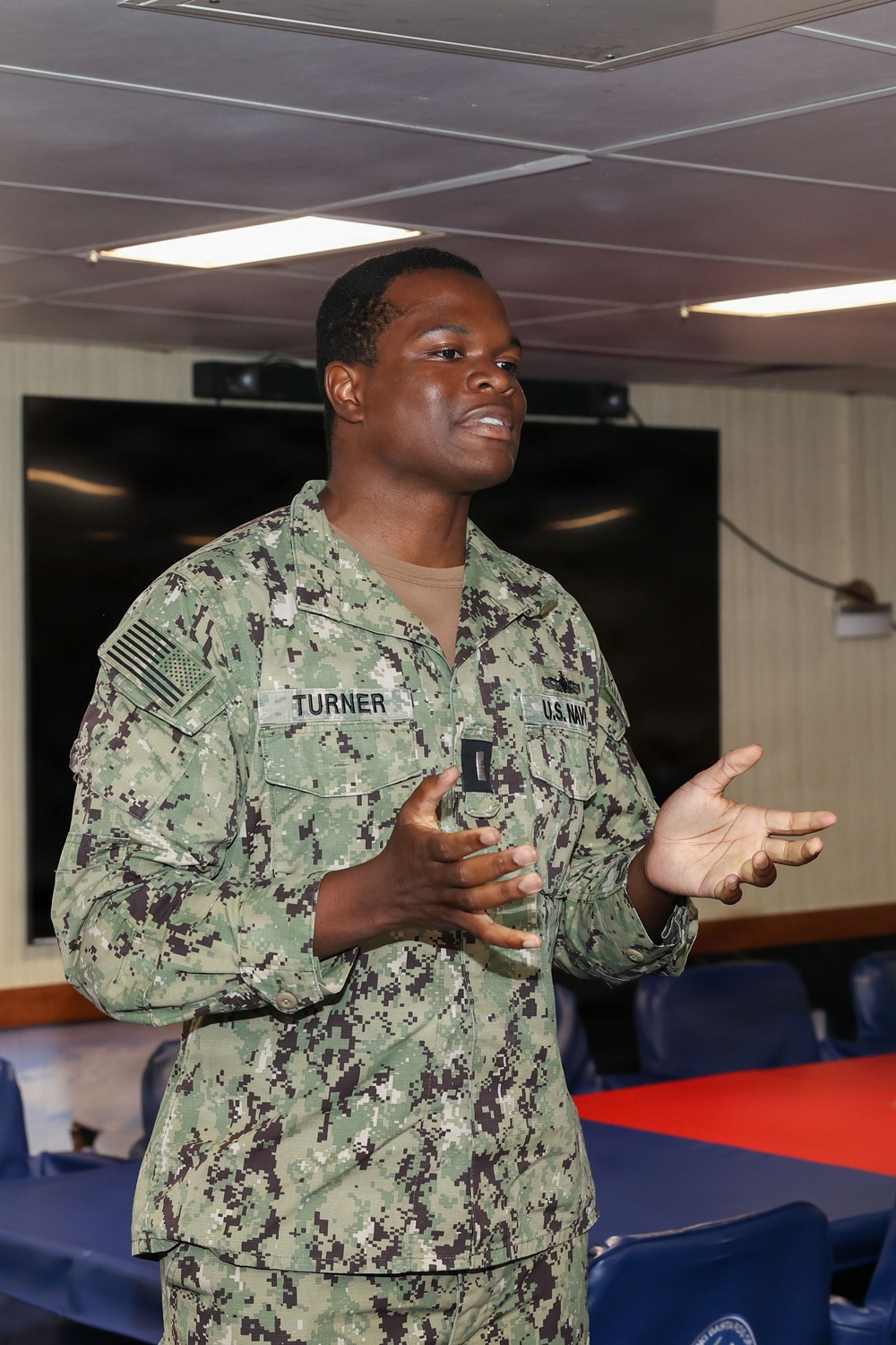 A U.S. Navy service member named Turner is speaking or presenting in what appears to be a briefing room or meeting space aboard a naval vessel or installation. The individual is wearing the Navy Working Uniform (NWU Type III) with a name tape reading 'TURNER' and a 'U.S. NAVY' tape, along with a U.S. flag patch on the left sleeve. Their hand gestures suggest active communication or instruction.