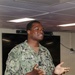 A U.S. Navy service member named Turner is speaking or presenting in what appears to be a briefing room or meeting space aboard a naval vessel or installation. The individual is wearing the Navy Working Uniform (NWU Type III) with a name tape reading 'TURNER' and a 'U.S. NAVY' tape, along with a U.S. flag patch on the left sleeve. Their hand gestures suggest active communication or instruction.