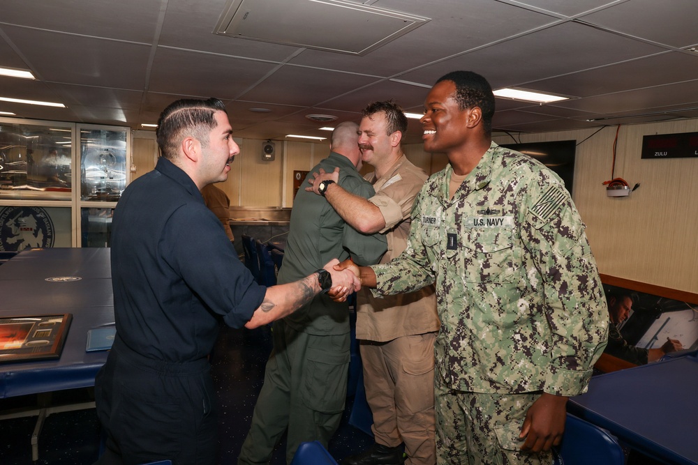 A U.S. Navy sailor in NWU Type III camouflage uniform is shaking hands with a man in dark civilian or working attire inside what appears to be a naval vessel's briefing or conference room. In the background, two other individuals in military uniforms are also greeting each other. The setting suggests a formal or semi-formal meet-and-greet or recognition event aboard a ship.
