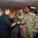 A U.S. Navy sailor in NWU Type III camouflage uniform is shaking hands with a man in dark civilian or working attire inside what appears to be a naval vessel's briefing or conference room. In the background, two other individuals in military uniforms are also greeting each other. The setting suggests a formal or semi-formal meet-and-greet or recognition event aboard a ship.