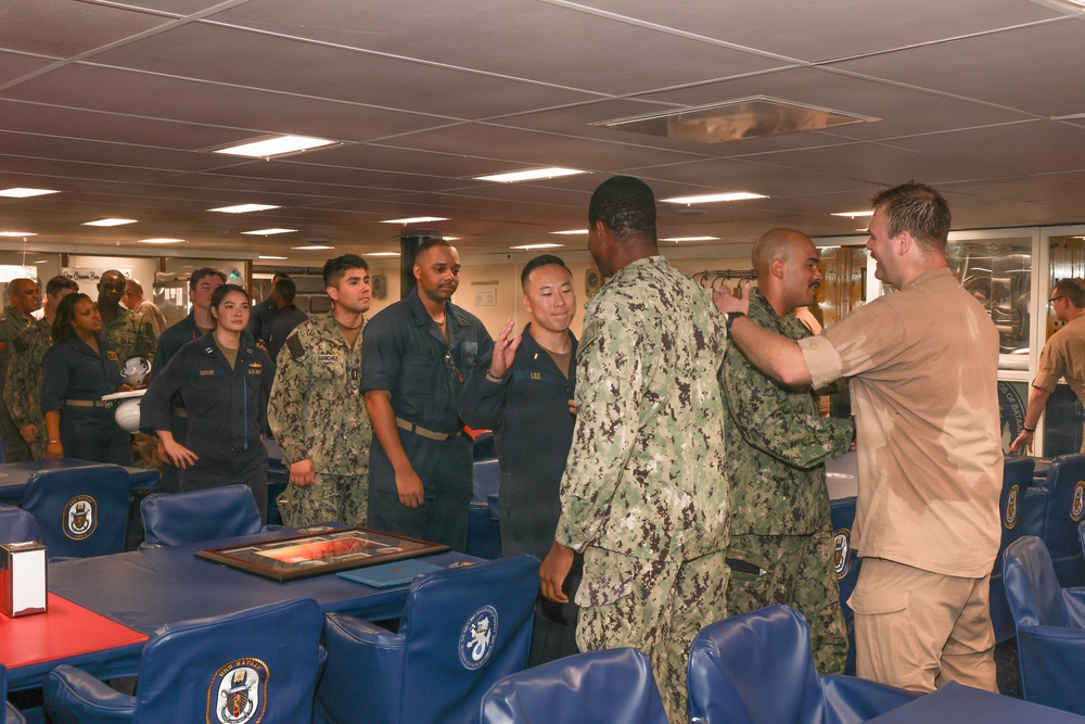 A group of U.S. Navy personnel are gathered in what appears to be a ship's mess hall or wardroom during what looks like a military ceremony or promotion event. Several sailors in Navy Working Uniforms (NWU) and khaki uniforms are interacting, with one individual appearing to have insignia or devices pinned to their uniform. Blue chairs bearing naval vessel insignia are visible throughout the room.