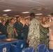 A group of U.S. Navy personnel are gathered in what appears to be a ship's mess hall or wardroom during what looks like a military ceremony or promotion event. Several sailors in Navy Working Uniforms (NWU) and khaki uniforms are interacting, with one individual appearing to have insignia or devices pinned to their uniform. Blue chairs bearing naval vessel insignia are visible throughout the room.