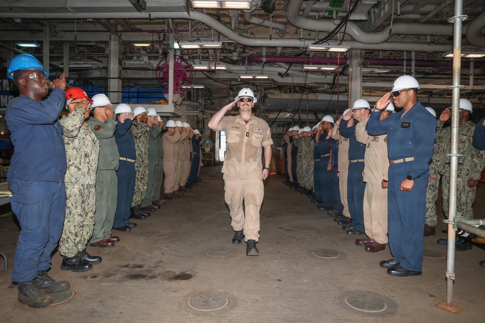 A U.S. Navy military ceremony is taking place inside what appears to be a ship's hangar bay or industrial deck space. A central figure in tan/khaki uniform wearing a white hard hat and sunglasses is walking through a corridor formed by two rows of saluting personnel. This appears to be a sideboy ceremony or a formal military honors passage, likely a change of command or departure ceremony.