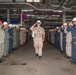 A U.S. Navy military ceremony is taking place inside what appears to be a ship's hangar bay or industrial deck space. A central figure in tan/khaki uniform wearing a white hard hat and sunglasses is walking through a corridor formed by two rows of saluting personnel. This appears to be a sideboy ceremony or a formal military honors passage, likely a change of command or departure ceremony.