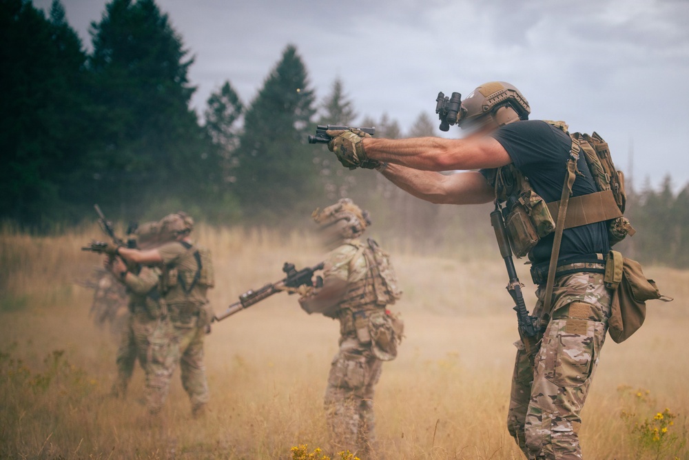 Three armed individuals in military-style tactical gear are positioned in an open field with dry grass, aiming firearms in various directions. The foreground figure wears a tactical helmet with mounted optics, a plate carrier, and multicam camouflage pants. Dust or smoke is visible in the background, suggesting an active training exercise or staged scenario.