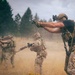 Three armed individuals in military-style tactical gear are positioned in an open field with dry grass, aiming firearms in various directions. The foreground figure wears a tactical helmet with mounted optics, a plate carrier, and multicam camouflage pants. Dust or smoke is visible in the background, suggesting an active training exercise or staged scenario.