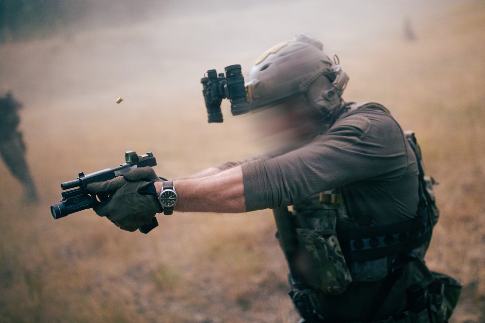 A tactical operator in full combat gear is firing a pistol equipped with a weapon light and red dot optic, with a spent brass casing visibly ejecting from the firearm. The individual is wearing a ballistic helmet with night vision device mount, multicam plate carrier, and tactical gloves. The setting appears to be an outdoor field environment with dust or smoke in the background.