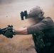 A tactical operator in full combat gear is firing a pistol equipped with a weapon light and red dot optic, with a spent brass casing visibly ejecting from the firearm. The individual is wearing a ballistic helmet with night vision device mount, multicam plate carrier, and tactical gloves. The setting appears to be an outdoor field environment with dust or smoke in the background.