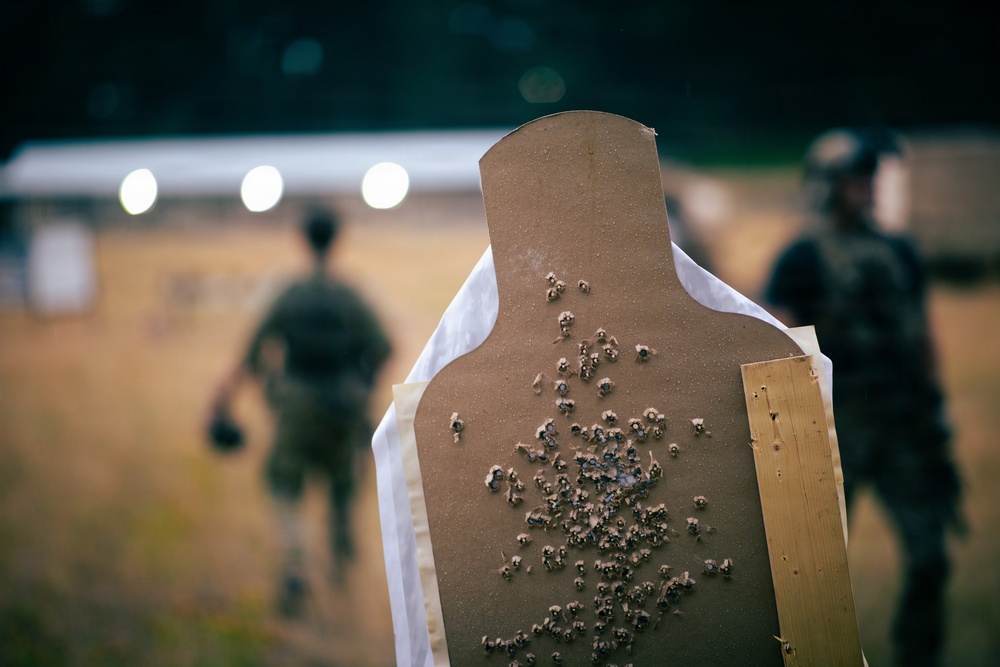 A close-up shot of a heavily perforated cardboard shooting target at what appears to be an outdoor firearms training range. The target shows a dense cluster of bullet holes concentrated in the center-mass area of the silhouette. Two individuals in tactical gear are visible in the blurred background.