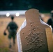 A close-up shot of a heavily perforated cardboard shooting target at what appears to be an outdoor firearms training range. The target shows a dense cluster of bullet holes concentrated in the center-mass area of the silhouette. Two individuals in tactical gear are visible in the blurred background.