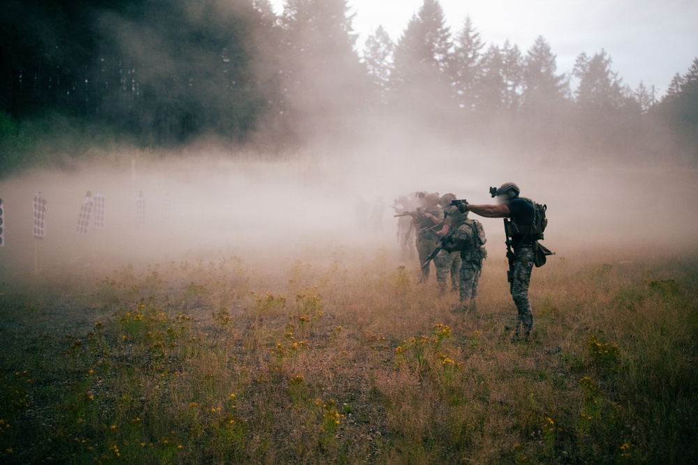 The image depicts what appears to be a military or tactical training exercise taking place in an open field surrounded by coniferous trees. A group of armed personnel in camouflage gear are advancing in a line formation through heavy smoke or dust, with paper/cardboard silhouette targets visible to the left. An instructor or evaluator figure stands to the right, appearing to direct or observe the exercise.