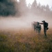 The image depicts what appears to be a military or tactical training exercise taking place in an open field surrounded by coniferous trees. A group of armed personnel in camouflage gear are advancing in a line formation through heavy smoke or dust, with paper/cardboard silhouette targets visible to the left. An instructor or evaluator figure stands to the right, appearing to direct or observe the exercise.
