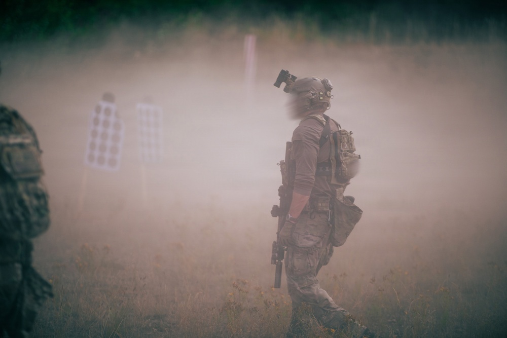 A heavily equipped tactical operator moves through a smoke-filled outdoor training range. The scene shows shooting targets visible in the background through the haze, suggesting an active live-fire or training exercise. The environment is an open grassy field with tree line visible in the far background.