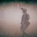A heavily equipped tactical operator moves through a smoke-filled outdoor training range. The scene shows shooting targets visible in the background through the haze, suggesting an active live-fire or training exercise. The environment is an open grassy field with tree line visible in the far background.