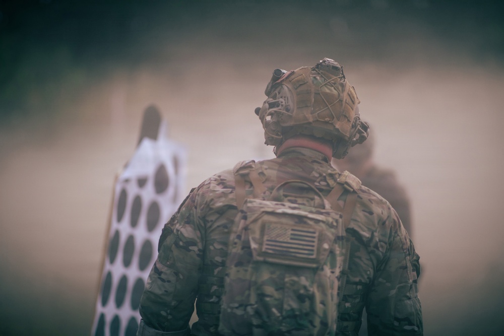 A U.S. military soldier is seen from behind, wearing full combat gear including a multicam camouflage uniform, tactical helmet with mounted equipment, and a backpack. The soldier appears to be on a firing range or training area, facing shooting targets partially obscured by dust or smoke. An American flag patch is visible on the soldier's right shoulder.