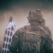 A U.S. military soldier is seen from behind, wearing full combat gear including a multicam camouflage uniform, tactical helmet with mounted equipment, and a backpack. The soldier appears to be on a firing range or training area, facing shooting targets partially obscured by dust or smoke. An American flag patch is visible on the soldier's right shoulder.