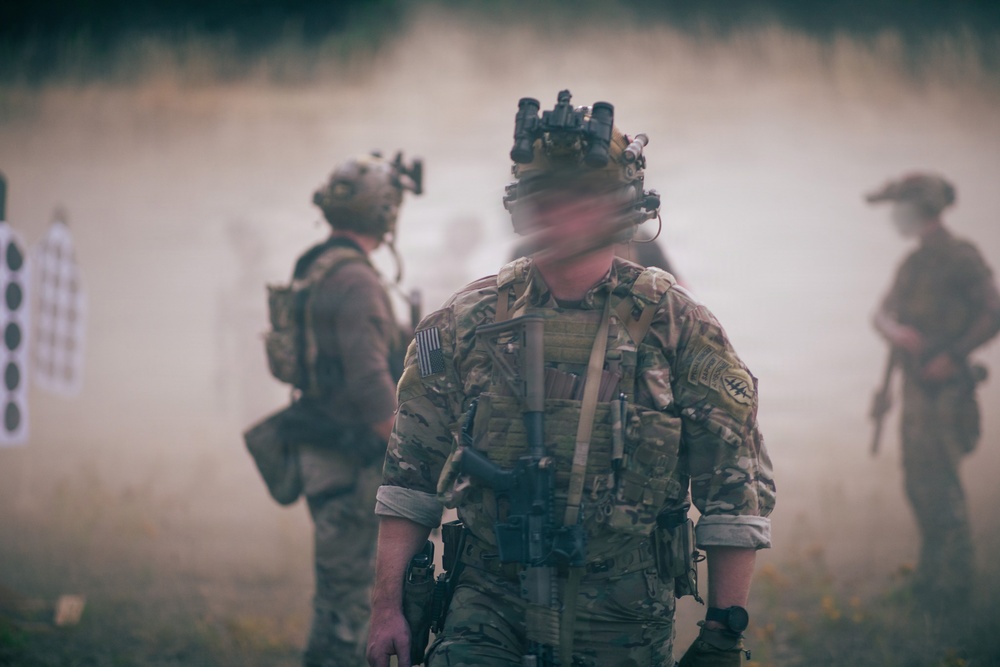 The image depicts military personnel in full tactical gear operating in a smoky outdoor environment, likely during a training exercise. At least three individuals are visible, with the foreground subject wearing multicam camouflage, a tactical vest, night vision device mount on helmet, and carrying a sidearm. Shooting targets are visible in the background left, strongly suggesting a live-fire or tactical training range scenario.