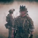 The image depicts military personnel in full tactical gear operating in a smoky outdoor environment, likely during a training exercise. At least three individuals are visible, with the foreground subject wearing multicam camouflage, a tactical vest, night vision device mount on helmet, and carrying a sidearm. Shooting targets are visible in the background left, strongly suggesting a live-fire or tactical training range scenario.