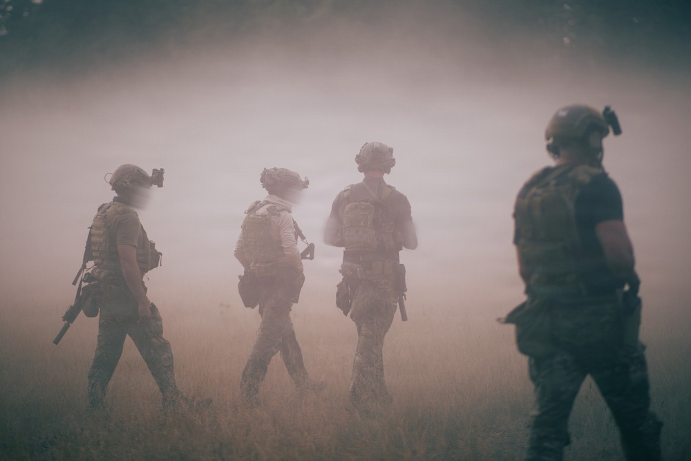 Four armed military personnel in full tactical gear are moving through a heavily smoke or dust-filled open field. The figures are silhouetted against a dense haze, obscuring background details. The scene depicts what appears to be a tactical patrol or training exercise in low-visibility conditions.