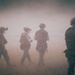 Four armed military personnel in full tactical gear are moving through a heavily smoke or dust-filled open field. The figures are silhouetted against a dense haze, obscuring background details. The scene depicts what appears to be a tactical patrol or training exercise in low-visibility conditions.