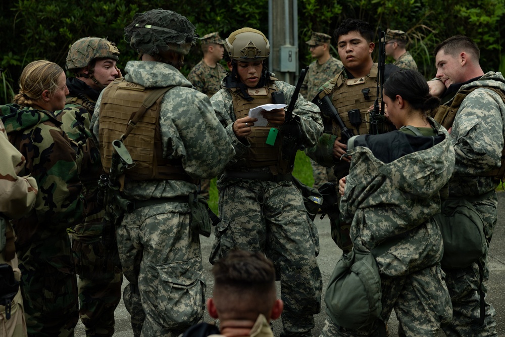 A group of military personnel in various camouflage uniforms are gathered in what appears to be a briefing or planning huddle outdoors. One central figure wearing a tactical helmet and plate carrier vest is reading from a document, suggesting a mission briefing or after-action review. The group includes both male and female service members carrying rifles and wearing full combat gear.
