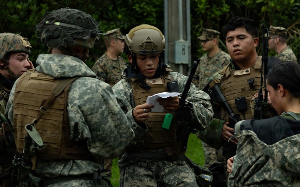 A group of military personnel in various camouflage uniforms are gathered in what appears to be a briefing or planning huddle outdoors. One central figure wearing a tactical helmet and plate carrier vest is reading from a document, suggesting a mission briefing or after-action review. The group includes both male and female service members carrying rifles and wearing full combat gear.