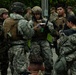 A group of military personnel in various camouflage uniforms are gathered in what appears to be a briefing or planning huddle outdoors. One central figure wearing a tactical helmet and plate carrier vest is reading from a document, suggesting a mission briefing or after-action review. The group includes both male and female service members carrying rifles and wearing full combat gear.