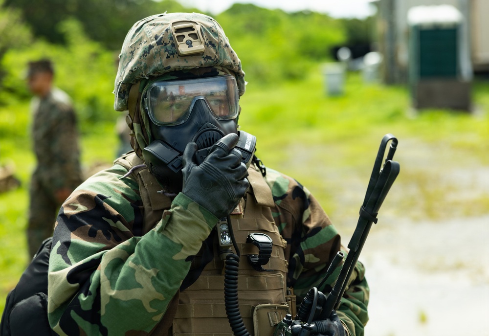 A military service member in full CBRN (Chemical, Biological, Radiological, Nuclear) protective gear is pictured in an outdoor setting. The individual is wearing a gas mask, digital camouflage helmet, woodland camouflage uniform, tactical vest, and black gloves, and appears to be adjusting or securing their gas mask. Two additional personnel in camouflage uniforms are visible in the blurred background.