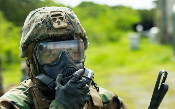 A military service member in full CBRN (Chemical, Biological, Radiological, Nuclear) protective gear is pictured in an outdoor setting. The individual is wearing a gas mask, digital camouflage helmet, woodland camouflage uniform, tactical vest, and black gloves, and appears to be adjusting or securing their gas mask. Two additional personnel in camouflage uniforms are visible in the blurred background.