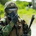 A military service member in full CBRN (Chemical, Biological, Radiological, Nuclear) protective gear is pictured in an outdoor setting. The individual is wearing a gas mask, digital camouflage helmet, woodland camouflage uniform, tactical vest, and black gloves, and appears to be adjusting or securing their gas mask. Two additional personnel in camouflage uniforms are visible in the blurred background.
