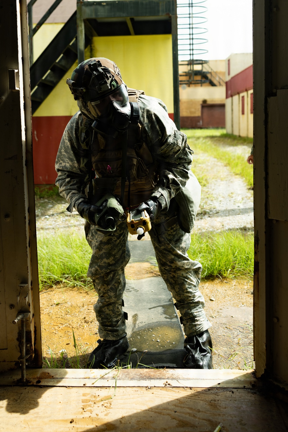 A soldier in full CBRN (Chemical, Biological, Radiological, Nuclear) protective gear stands in a doorway of what appears to be a military training facility. The individual is wearing US Army ACU (Army Combat Uniform) pattern camouflage, a gas mask, tactical helmet with night vision mount, and black rubber overboots. The soldier appears to be holding a detection or sampling device and looking downward, suggesting active CBRN reconnaissance or decontamination training.