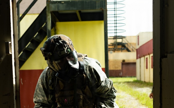 A soldier in full CBRN (Chemical, Biological, Radiological, Nuclear) protective gear stands in a doorway of what appears to be a military training facility. The individual is wearing US Army ACU (Army Combat Uniform) pattern camouflage, a gas mask, tactical helmet with night vision mount, and black rubber overboots. The soldier appears to be holding a detection or sampling device and looking downward, suggesting active CBRN reconnaissance or decontamination training.