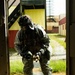 A soldier in full CBRN (Chemical, Biological, Radiological, Nuclear) protective gear stands in a doorway of what appears to be a military training facility. The individual is wearing US Army ACU (Army Combat Uniform) pattern camouflage, a gas mask, tactical helmet with night vision mount, and black rubber overboots. The soldier appears to be holding a detection or sampling device and looking downward, suggesting active CBRN reconnaissance or decontamination training.